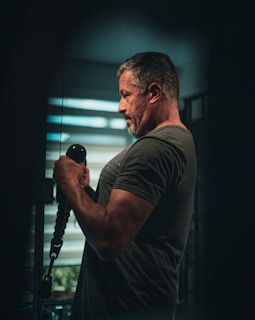 A man with short hair and a beard is performing a bicep curl exercise using a cable machine. He is wearing a dark t-shirt, standing in a dimly lit room with a focused expression. The background shows slatted blinds allowing for some light to pass through.
