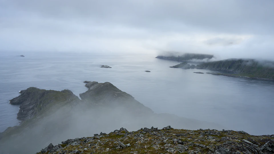The image depicts a serene coastal landscape with a rocky cliff overlooking a vast body of water. The sky above is filled with clouds, creating an overcast atmosphere that adds to the overall tranquility of the scene. In the distance, several small islands can be seen floating on the water's surface.
