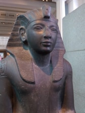 An ancient Egyptian statue depicting a figure wearing a nemes headdress with uraeus, typical of pharaohs. The statue is carved from dark stone, with finely detailed facial features and a serene expression. The background includes museum interior elements such as pillars and architectural details.