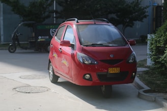 A red compact car is parked on the side of a road near some bushes, with a three-wheeled vehicle in the background. The car has a yellow license plate and decorative decals on the side. The scene has a mix of natural and urban elements.