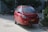 A red compact car is parked on the side of a road near some bushes, with a three-wheeled vehicle in the background. The car has a yellow license plate and decorative decals on the side. The scene has a mix of natural and urban elements.