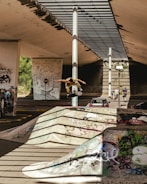 In an urban skate park, a skateboarder performs a mid-air trick against a backdrop of vibrant graffiti on ramps and pillars. The area is shaded by an overhead structure with a combination of concrete and metal. Vehicles and pedestrians are visible in the background, contributing to the lively atmosphere of the scene.