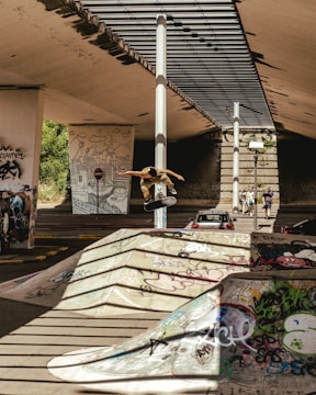 In an urban skate park, a skateboarder performs a mid-air trick against a backdrop of vibrant graffiti on ramps and pillars. The area is shaded by an overhead structure with a combination of concrete and metal. Vehicles and pedestrians are visible in the background, contributing to the lively atmosphere of the scene.