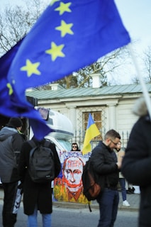 A public protest scene includes several individuals standing near a painted sign with a striking image and text. Flags with European Union and Ukrainian colors are prominently displayed, and a building with classical architecture forms the backdrop.