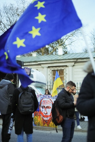 A public protest scene includes several individuals standing near a painted sign with a striking image and text. Flags with European Union and Ukrainian colors are prominently displayed, and a building with classical architecture forms the backdrop.