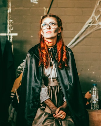 A person wearing glasses with red hair styled in curls, wearing a black cape and a Victorian-style dress. The face is painted with makeup resembling a Halloween or gothic character, featuring pale skin and shadowed cheeks. The background includes cobwebs and a brick wall, giving a spooky atmosphere.
