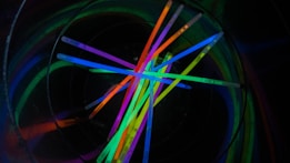 A collection of vibrant glow sticks arranged in a crisscross pattern inside a dark, circular container. The glow sticks emit a bright, multi-colored light with shades of green, blue, pink, orange, and purple, contrasting against the black background.