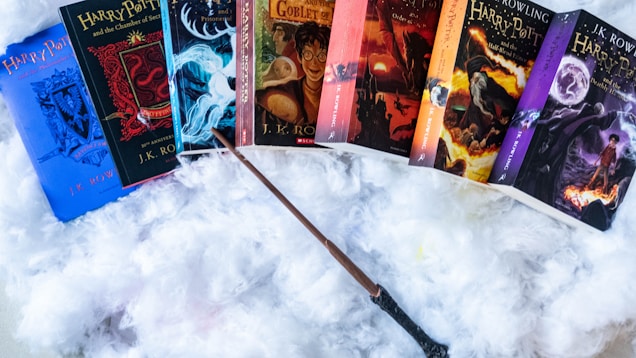 A collection of seven books from the Harry Potter series is displayed on a fluffy white surface. Each book has a colorful and distinct cover design. A wand is placed in front of the books, creating a magical theme.