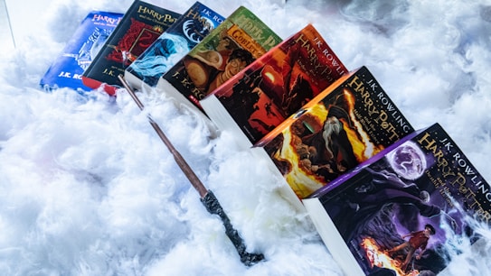 A collection of Harry Potter books are arranged in a fan-like pattern on a soft, fluffy background. A wand is diagonally placed across the lower part of the books, adding a magical element.