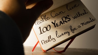A hand holding a notebook open to a page with handwritten text that reads: 'People can live 100 years without really living for a minute.' The page appears to be well-lit, while the surrounding area is slightly shadowed.