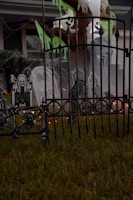 A spooky outdoor Halloween decoration featuring skull motifs on a wrought iron fence, tombstones with eerie designs, and ghostly figures hanging from a tree. Cobwebs enhance the haunted atmosphere, and there are faint orange lights glowing in the background.