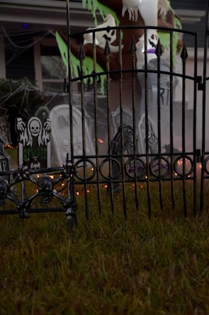 A spooky outdoor Halloween decoration featuring skull motifs on a wrought iron fence, tombstones with eerie designs, and ghostly figures hanging from a tree. Cobwebs enhance the haunted atmosphere, and there are faint orange lights glowing in the background.