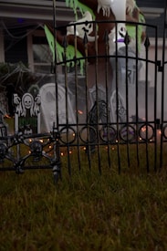 A spooky outdoor Halloween decoration featuring skull motifs on a wrought iron fence, tombstones with eerie designs, and ghostly figures hanging from a tree. Cobwebs enhance the haunted atmosphere, and there are faint orange lights glowing in the background.