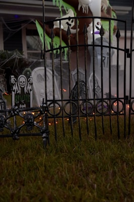 A spooky outdoor Halloween decoration featuring skull motifs on a wrought iron fence, tombstones with eerie designs, and ghostly figures hanging from a tree. Cobwebs enhance the haunted atmosphere, and there are faint orange lights glowing in the background.