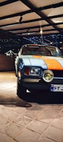 A classic car parked indoors, featuring a distinctive blue and orange paint job. The setting is well-lit with spotlights, and a brick wall is visible in the background. The car's headlights are prominent, and the area exudes a vintage aesthetic.