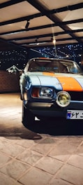 A classic car parked indoors, featuring a distinctive blue and orange paint job. The setting is well-lit with spotlights, and a brick wall is visible in the background. The car's headlights are prominent, and the area exudes a vintage aesthetic.