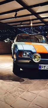 A classic car parked indoors, featuring a distinctive blue and orange paint job. The setting is well-lit with spotlights, and a brick wall is visible in the background. The car's headlights are prominent, and the area exudes a vintage aesthetic.