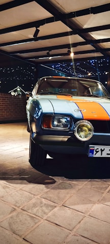 A classic car parked indoors, featuring a distinctive blue and orange paint job. The setting is well-lit with spotlights, and a brick wall is visible in the background. The car's headlights are prominent, and the area exudes a vintage aesthetic.