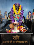 A statue of a deity is adorned with vibrant garlands of purple and yellow flowers. The black statue is situated on a platform with various offerings, including a coconut, a silver pot, and a small oil lamp. There is incense smoke wafting over the scene, and another garlanded idol can be seen in the background.