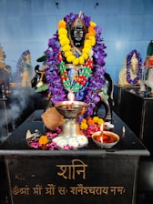 A statue of a deity is adorned with vibrant garlands of purple and yellow flowers. The black statue is situated on a platform with various offerings, including a coconut, a silver pot, and a small oil lamp. There is incense smoke wafting over the scene, and another garlanded idol can be seen in the background.