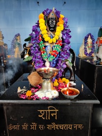 A statue of a deity is adorned with vibrant garlands of purple and yellow flowers. The black statue is situated on a platform with various offerings, including a coconut, a silver pot, and a small oil lamp. There is incense smoke wafting over the scene, and another garlanded idol can be seen in the background.