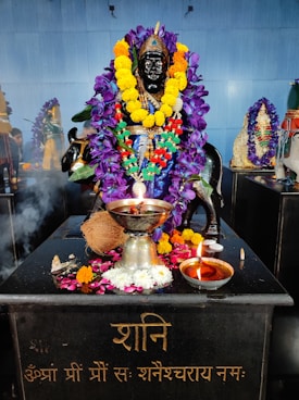 A statue of a deity is adorned with vibrant garlands of purple and yellow flowers. The black statue is situated on a platform with various offerings, including a coconut, a silver pot, and a small oil lamp. There is incense smoke wafting over the scene, and another garlanded idol can be seen in the background.