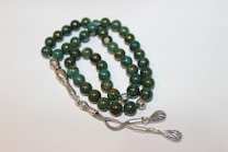 A strand of prayer beads with polished green stones, intricately designed silver separators, and decorative metallic tassels.