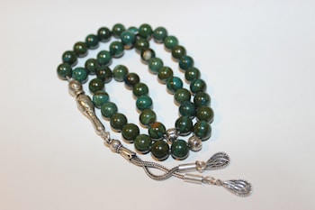 A strand of prayer beads with polished green stones, intricately designed silver separators, and decorative metallic tassels.
