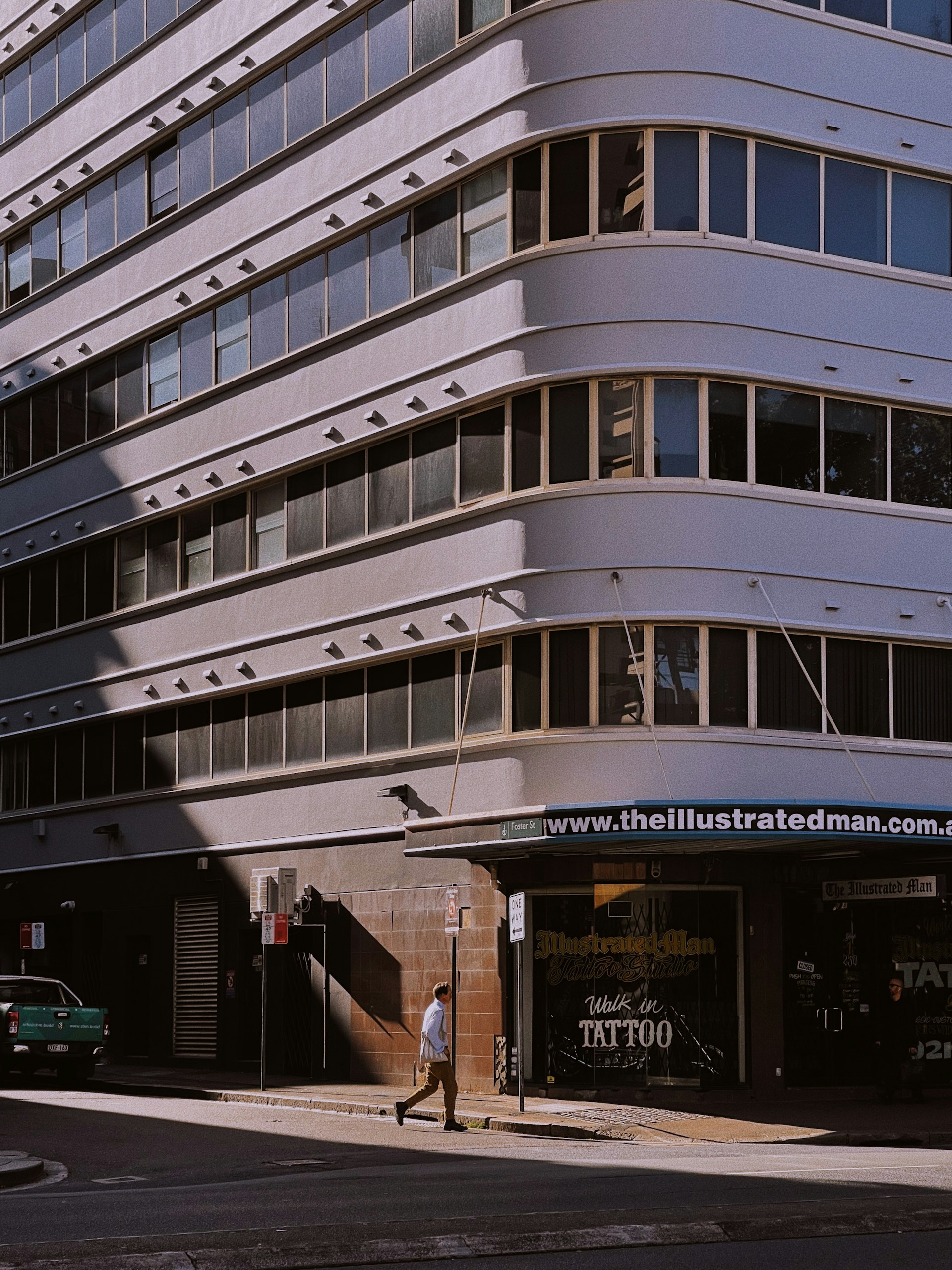 A modern building with curved lines and large glass windows is partially covered in shadows. A person is walking on the sidewalk past a tattoo studio with a sign that reads 'Walk in Tattoo'. The building's facade is white, and there is another parked vehicle along the street.