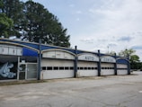 A vacant auto center with an old, worn-down facade featuring three large garage doors and bold white lettering at the top. There are some graffiti on the glass doors to the left. Tall trees are visible behind the building, and the surrounding area appears deserted with cracked concrete.
