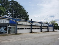 A vacant auto center with an old, worn-down facade featuring three large garage doors and bold white lettering at the top. There are some graffiti on the glass doors to the left. Tall trees are visible behind the building, and the surrounding area appears deserted with cracked concrete.