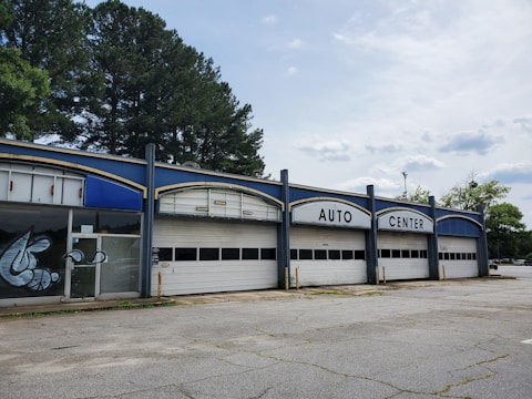 A vacant auto center with an old, worn-down facade featuring three large garage doors and bold white lettering at the top. There are some graffiti on the glass doors to the left. Tall trees are visible behind the building, and the surrounding area appears deserted with cracked concrete.