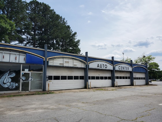 A vacant auto center with an old, worn-down facade featuring three large garage doors and bold white lettering at the top. There are some graffiti on the glass doors to the left. Tall trees are visible behind the building, and the surrounding area appears deserted with cracked concrete.