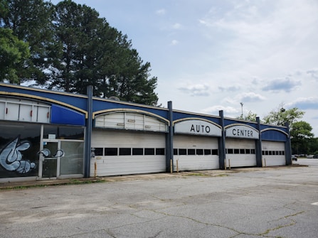 A vacant auto center with an old, worn-down facade featuring three large garage doors and bold white lettering at the top. There are some graffiti on the glass doors to the left. Tall trees are visible behind the building, and the surrounding area appears deserted with cracked concrete.