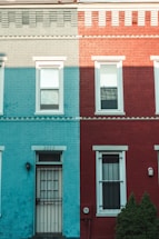 A building facade is painted in two distinct colors, turquoise on the left and dark red on the right. The structure features a door and windows with white trims, and a metal gate covers the door. The numbers 2222 are displayed above the door. A small bush is visible on the right near the building.