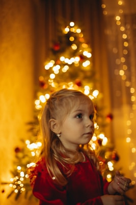 A young child with blonde hair is wearing a red dress. In the background, a Christmas tree adorned with warm white lights and red ornaments creates a festive atmosphere. The lighting is soft and warm, enhancing the cozy, holiday mood.