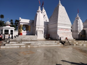 A white temple complex with multiple spires and Hindi text on the walls. People are standing near the entrance, and the area is surrounded by clear skies and palm trees in the background.