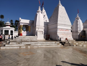 A white temple complex with multiple spires and Hindi text on the walls. People are standing near the entrance, and the area is surrounded by clear skies and palm trees in the background.