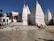 A white temple complex with multiple spires and Hindi text on the walls. People are standing near the entrance, and the area is surrounded by clear skies and palm trees in the background.