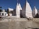 A white temple complex with multiple spires and Hindi text on the walls. People are standing near the entrance, and the area is surrounded by clear skies and palm trees in the background.