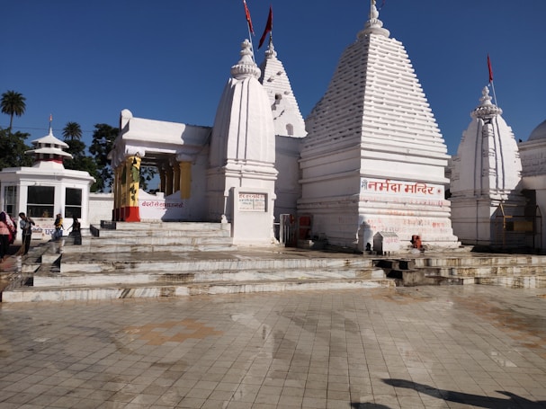 A white temple complex with multiple spires and Hindi text on the walls. People are standing near the entrance, and the area is surrounded by clear skies and palm trees in the background.