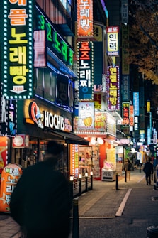 A vibrant city street at night filled with colorful neon signs in various languages. The scene includes a blurred silhouette of a person walking along the sidewalk, and several small shops with illuminated signage. The street is bustling with energy and the lighting casts a lively glow across the environment.