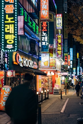 A vibrant city street at night filled with colorful neon signs in various languages. The scene includes a blurred silhouette of a person walking along the sidewalk, and several small shops with illuminated signage. The street is bustling with energy and the lighting casts a lively glow across the environment.