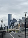 A cityscape featuring a blend of modern skyscrapers and historic architecture under an overcast sky. Street lamps and benches line the empty pathway, leading towards tram stops, while people can be seen in the distance near the transport area.