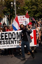 A group of people are participating in a protest. They are holding a large banner that reads 'FREEDOM ESSENTIAL @ QUEENS PARK EVERY SATURDAY'. One of the people at the front of the group is speaking through a megaphone. A sign in the background reads 'I ♥ FREE SPEECH,' and there is a visible Canadian flag on the banner. Some people are holding flags and wearing hats, and the scene takes place outdoors with trees in the background.