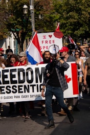 A group of people are participating in a protest. They are holding a large banner that reads 'FREEDOM ESSENTIAL @ QUEENS PARK EVERY SATURDAY'. One of the people at the front of the group is speaking through a megaphone. A sign in the background reads 'I ♥ FREE SPEECH,' and there is a visible Canadian flag on the banner. Some people are holding flags and wearing hats, and the scene takes place outdoors with trees in the background.