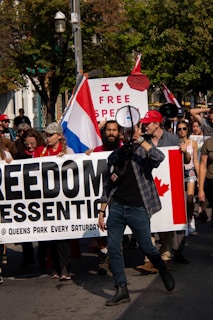 A group of people are participating in a protest. They are holding a large banner that reads 'FREEDOM ESSENTIAL @ QUEENS PARK EVERY SATURDAY'. One of the people at the front of the group is speaking through a megaphone. A sign in the background reads 'I ♥ FREE SPEECH,' and there is a visible Canadian flag on the banner. Some people are holding flags and wearing hats, and the scene takes place outdoors with trees in the background.