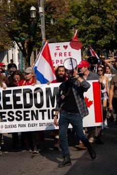 A group of people are participating in a protest. They are holding a large banner that reads 'FREEDOM ESSENTIAL @ QUEENS PARK EVERY SATURDAY'. One of the people at the front of the group is speaking through a megaphone. A sign in the background reads 'I ♥ FREE SPEECH,' and there is a visible Canadian flag on the banner. Some people are holding flags and wearing hats, and the scene takes place outdoors with trees in the background.