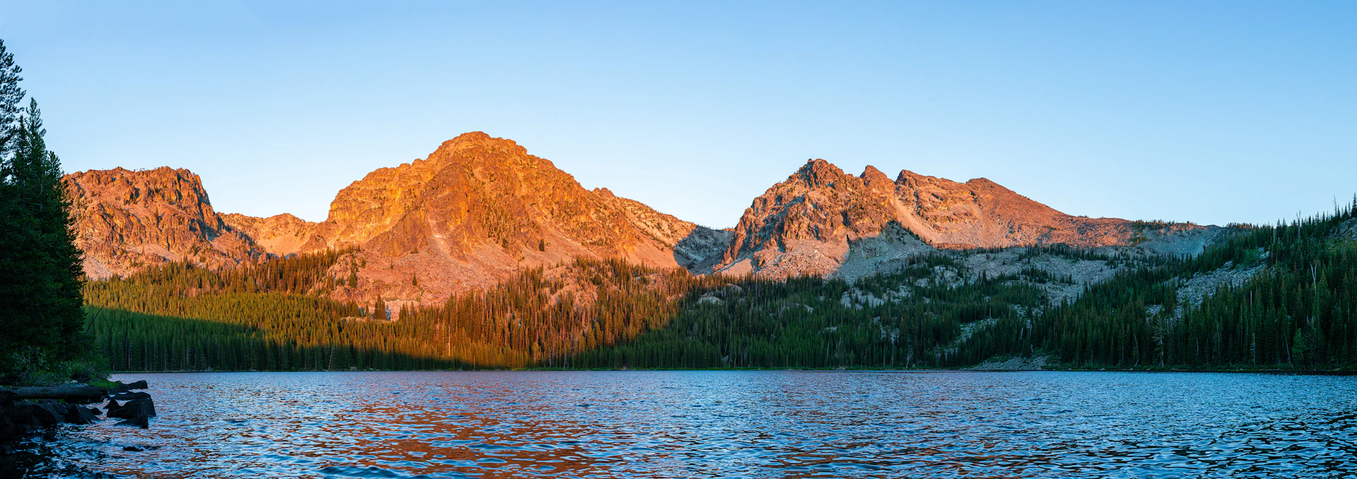 A panoramic view of a rugged mountain range at sunset, with vibrant orange and purple hues reflecting off a calm lake below.