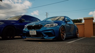A blue sports car with customized details is parked next to a larger blue vehicle. The car has a personalized license plate and distinctive bronze-colored rims. The setting appears to be an outdoor parking lot with clear skies and scattered clouds.